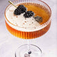 A coupe glass brimming with a bubbly amber cocktail, elegantly garnished with two blackberries on a pick, captures the stunning style showcased in Cocktails Illustrated by Penguin Random House—perfect inspiration for your next cocktail creation or to elevate your bartending repertoire.