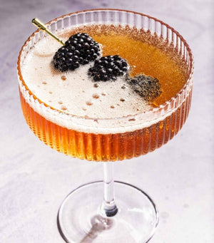 A coupe glass brimming with a bubbly amber cocktail, elegantly garnished with two blackberries on a pick, captures the stunning style showcased in Cocktails Illustrated by Penguin Random House—perfect inspiration for your next cocktail creation or to elevate your bartending repertoire.