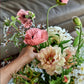 A hand arranges a bouquet of assorted flowers—pink poppies and cream peonies among them—in the elegant, signature style showcased in Home in Bloom by Penguin Random House, with a softly blurred couch in the background.