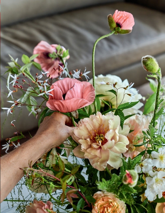 A hand arranges a bouquet of assorted flowers—pink poppies and cream peonies among them—in the elegant, signature style showcased in Home in Bloom by Penguin Random House, with a softly blurred couch in the background.