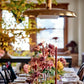 A dining table is set with floral arrangements inspired by Home in Bloom from Penguin Random House and tall candles, with plates and glassware displayed under a brass pendant light in a room adorned with autumnal decor.