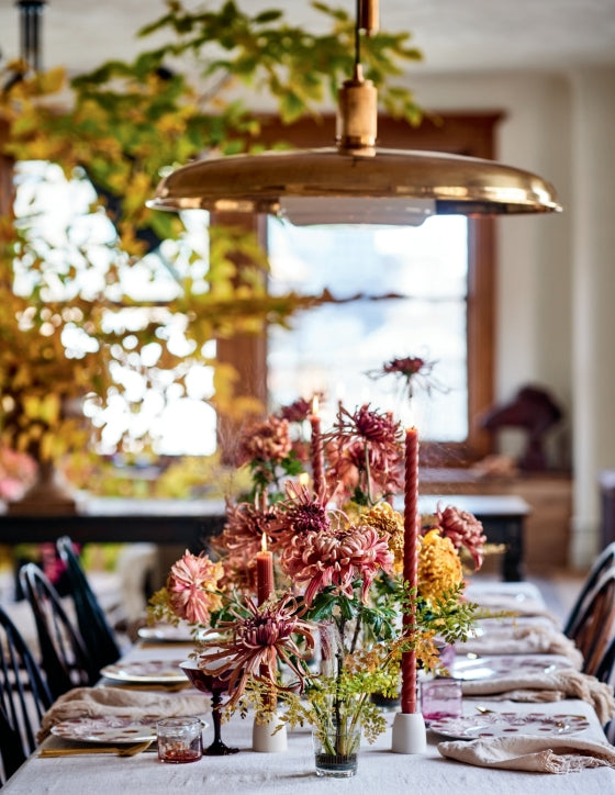 A dining table is set with floral arrangements inspired by Home in Bloom from Penguin Random House and tall candles, with plates and glassware displayed under a brass pendant light in a room adorned with autumnal decor.