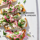 Flatbread slices with cheese, pickled onions, fresh herbs, and green onions are arranged on a white board beside the text “Boards & Spreads,” inspired by the product *Boards & Spreads* from Penguin Random House—perfect for sparking ideas for your next appetizer spread or recipe board.