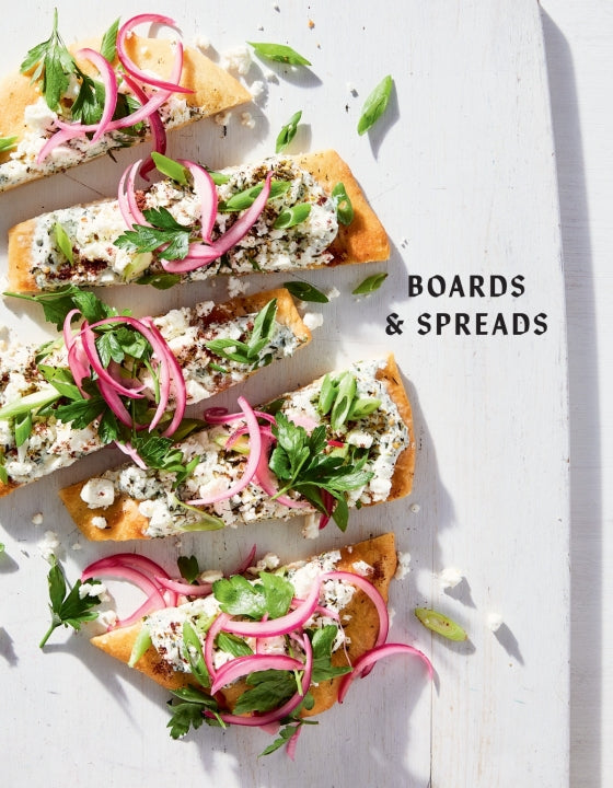 Flatbread slices with cheese, pickled onions, fresh herbs, and green onions are arranged on a white board beside the text “Boards & Spreads,” inspired by the product *Boards & Spreads* from Penguin Random House—perfect for sparking ideas for your next appetizer spread or recipe board.