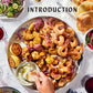 A hand dips bread into a bowl of spread on a platter featuring grilled shrimp, chicken, onions, lemon, and sauce—an enticing array inspired by the artful presentations found in Boards & Spreads from Penguin Random House. Surrounded by salad, extra bread, and glasses of red wine atop a white tablecloth, this inviting scene is perfect for sharing as part of generous snack platters or elegant recipe boards.