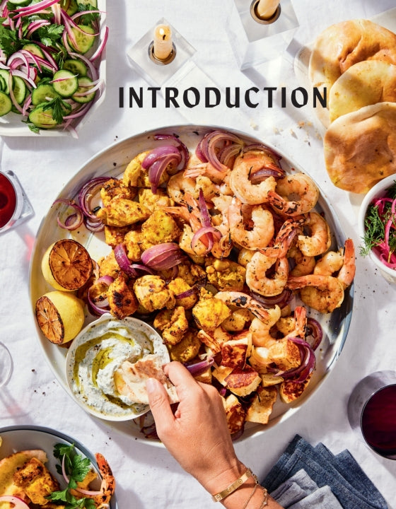 A hand dips bread into a bowl of spread on a platter featuring grilled shrimp, chicken, onions, lemon, and sauce—an enticing array inspired by the artful presentations found in Boards & Spreads from Penguin Random House. Surrounded by salad, extra bread, and glasses of red wine atop a white tablecloth, this inviting scene is perfect for sharing as part of generous snack platters or elegant recipe boards.