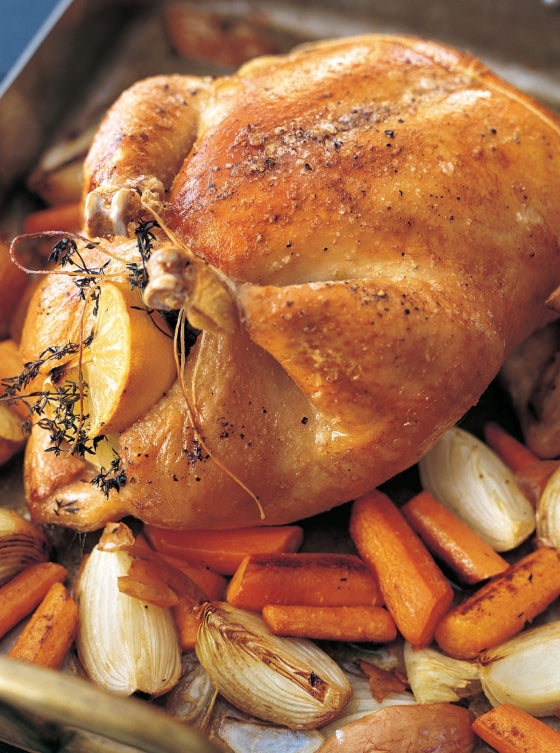 A whole roasted chicken with golden-brown skin, inspired by Barefoot Contessa At Home from Penguin Random House, stuffed with lemon and herbs and nestled on a bed of roasted carrots and onions in a baking pan—a classic East Hampton dinner.