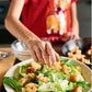 Wearing a red shirt with a cartoon dog illustration, a person sprinkles croutons onto a Caesar salad with romaine lettuce, recreating one of the easy recipes from “Kid in the Kitchen” by Penguin Random House.
