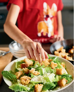 Wearing a red shirt with a cartoon dog illustration, a person sprinkles croutons onto a Caesar salad with romaine lettuce, recreating one of the easy recipes from “Kid in the Kitchen” by Penguin Random House.