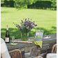 From Barefoot Contessa Foolproof by Penguin Random House: A vase brimming with purple flowers, a wine bottle accompanied by two glasses, and a water pitcher rest atop a stone ledge—set against outdoor seating and a lush green lawn—creating the perfect inviting atmosphere to savor dishes like Slow-Roasted Filet of Beef.