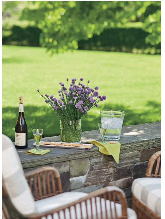 From Barefoot Contessa Foolproof by Penguin Random House: A vase brimming with purple flowers, a wine bottle accompanied by two glasses, and a water pitcher rest atop a stone ledge—set against outdoor seating and a lush green lawn—creating the perfect inviting atmosphere to savor dishes like Slow-Roasted Filet of Beef.