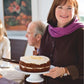 A woman wearing a purple scarf and smiling indoors presents a frosted cake on a stand, while two people seated at a table in the background enjoy dishes such as Slow-Roasted Filet of Beef and Hot Smoked Salmon, all inspired by recipes from Barefoot Contessa Foolproof by Penguin Random House.