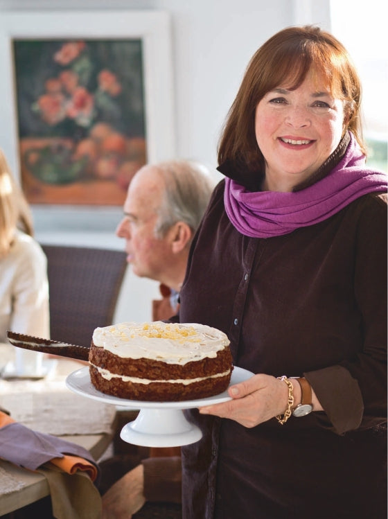 A woman wearing a purple scarf and smiling indoors presents a frosted cake on a stand, while two people seated at a table in the background enjoy dishes such as Slow-Roasted Filet of Beef and Hot Smoked Salmon, all inspired by recipes from Barefoot Contessa Foolproof by Penguin Random House.