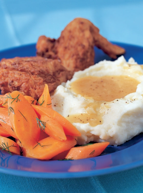 A comforting blue plate featuring fried chicken, mashed potatoes with savory gravy, and dill-garnished sliced carrots—perfectly capturing the inviting spirit of home-cooked meals celebrated in Barefoot Contessa Family Style from Penguin Random House.
