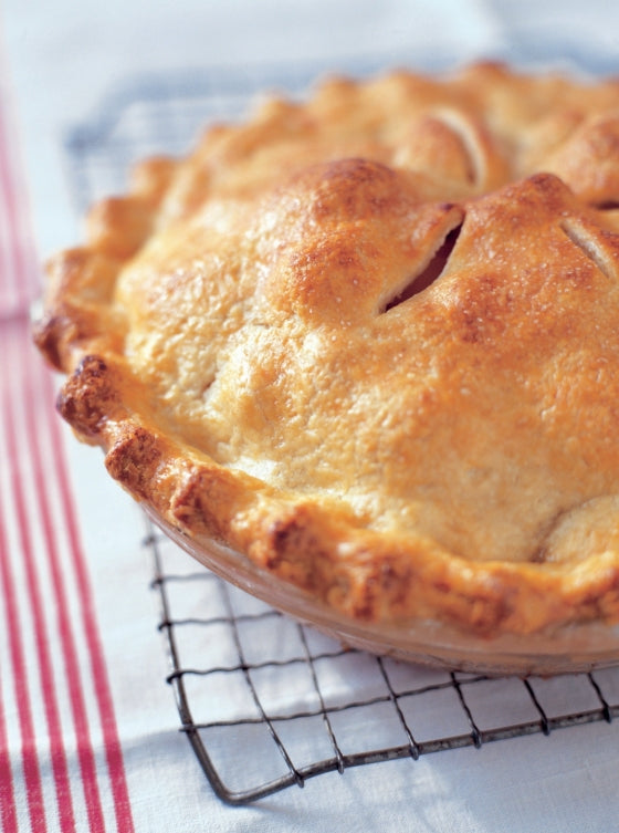 A golden-brown baked pie with a flaky crust, inspired by recipes from the “Barefoot Contessa Family Style” cookbook by Penguin Random House, cools on a wire rack over a white cloth with red stripes—an ideal treat for everyday cooking and family gatherings.