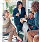 Four adults sit and stand on a porch, smiling and clinking glasses as they enjoy a casual outdoor gathering, sharing stories inspired by recipes from *Barefoot Contessa Family Style* by Penguin Random House—proof that everyday cooking can bring people together in the most joyful way.