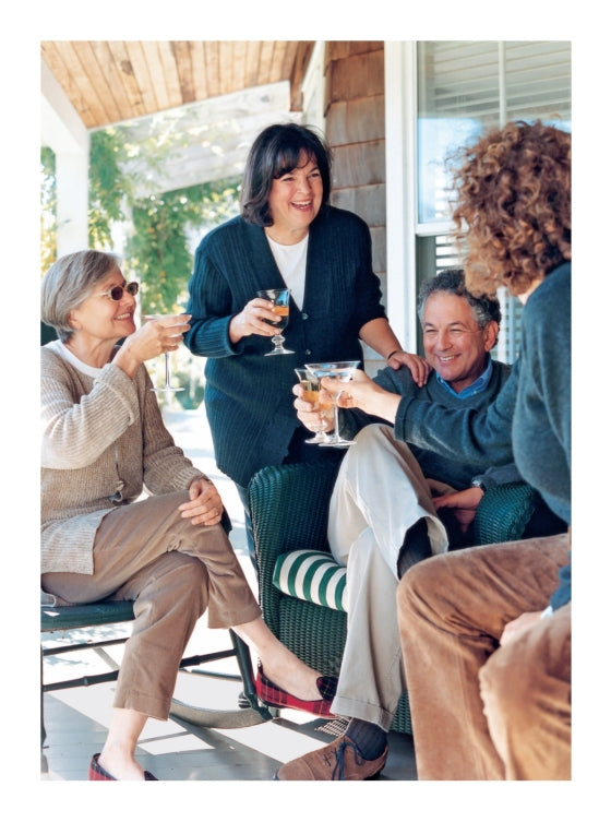Four adults sit and stand on a porch, smiling and clinking glasses as they enjoy a casual outdoor gathering, sharing stories inspired by recipes from *Barefoot Contessa Family Style* by Penguin Random House—proof that everyday cooking can bring people together in the most joyful way.