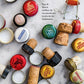 Assorted bottle caps and corks are scattered on a marble surface beside French text reading, “Pour B: Que le meilleur de nos passés soit le pire de nos lendemains,” capturing the spirit of a French apéritif experience as celebrated in Penguin Random House’s Apéritif.