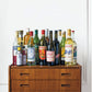 Several bottles featured in Apéritif by Penguin Random House—perfect for mixing classic French cocktails or savoring a traditional French apéritif with hors d’oeuvres—are displayed atop a wooden chest of drawers set against a simple white wall.