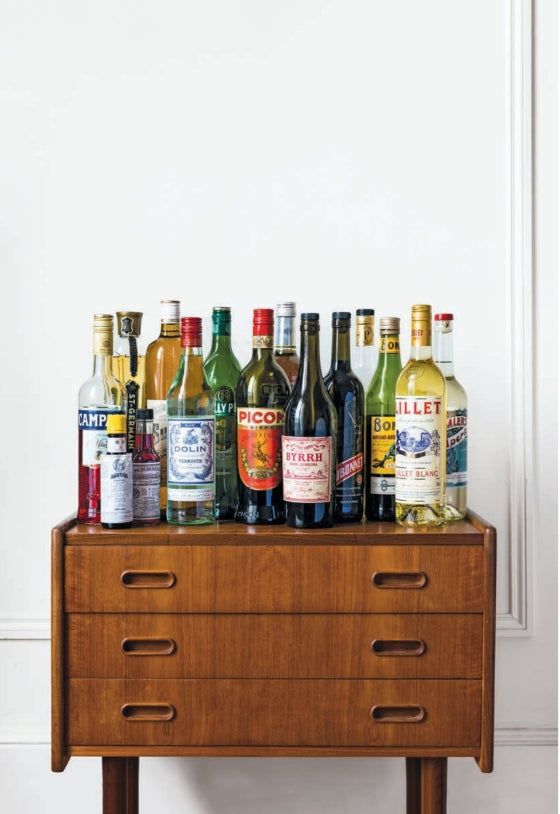 Several bottles featured in Apéritif by Penguin Random House—perfect for mixing classic French cocktails or savoring a traditional French apéritif with hors d’oeuvres—are displayed atop a wooden chest of drawers set against a simple white wall.
