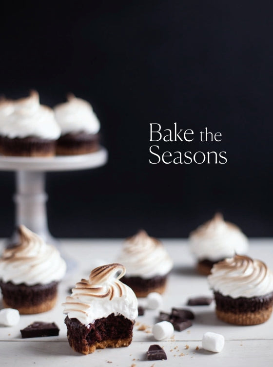 Swirled chocolate cupcakes are displayed on a stand and table, surrounded by marshmallows and chocolate pieces for a tempting comfort food scene. The cover features the title “Bake the Seasons” from Penguin Random House, evoking the heart of seasonal baking recipes against a dark backdrop.