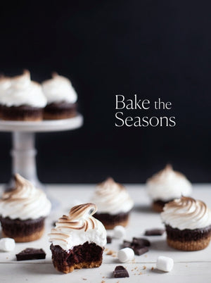 Swirled chocolate cupcakes are displayed on a stand and table, surrounded by marshmallows and chocolate pieces for a tempting comfort food scene. The cover features the title “Bake the Seasons” from Penguin Random House, evoking the heart of seasonal baking recipes against a dark backdrop.
