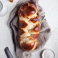 A copy of Bake the Seasons by Penguin Random House lies beside a braided loaf of challah bread on a linen cloth, accompanied by a small bowl of salt, a bread knife, and a honey dipper—inviting you to savor one of the ultimate comfort foods.