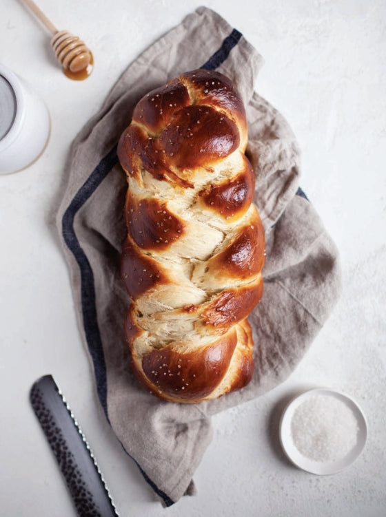 A copy of Bake the Seasons by Penguin Random House lies beside a braided loaf of challah bread on a linen cloth, accompanied by a small bowl of salt, a bread knife, and a honey dipper—inviting you to savor one of the ultimate comfort foods.