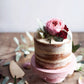 A petite semi-naked cake adorned with pink and red flowers rests on a pink cake stand atop a wooden table sprinkled with leaves and accompanied by a cake server, beautifully reflecting the spirit of *Bake the Seasons* by Penguin Random House and its comforting collection of seasonal baking recipes.