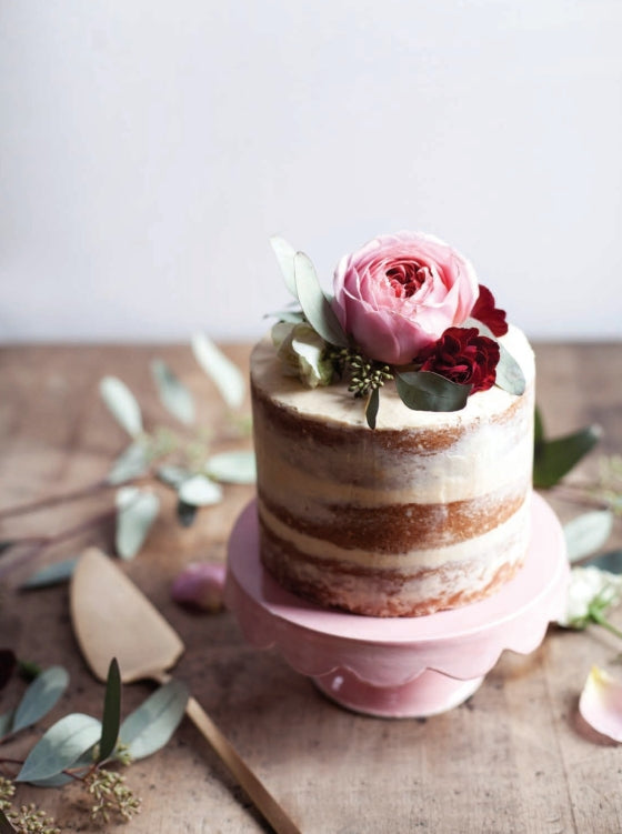 A petite semi-naked cake adorned with pink and red flowers rests on a pink cake stand atop a wooden table sprinkled with leaves and accompanied by a cake server, beautifully reflecting the spirit of *Bake the Seasons* by Penguin Random House and its comforting collection of seasonal baking recipes.