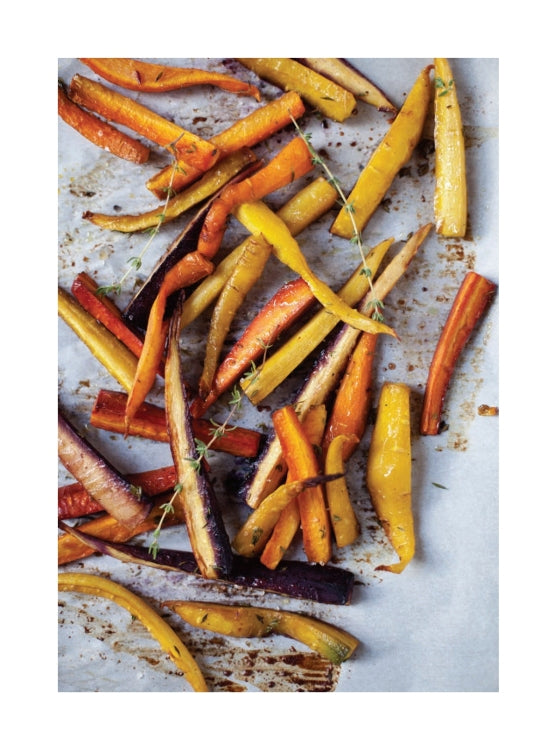Colorful roasted carrots, sliced into strips and scattered on a parchment-lined baking sheet with a few sprigs of fresh herbs—a vibrant dish perfect for fans of comfort food inspired by the freshest ingredients, just like the recipes found in *Bake the Seasons* from Penguin Random House.