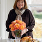 A woman stands indoors holding a vase of orange roses behind a table set with dishes, glassware, and a beautifully roasted chicken—the perfect setting to inspire easy dinner ideas or comforting recipes from Go-to Dinners by Penguin Random House.