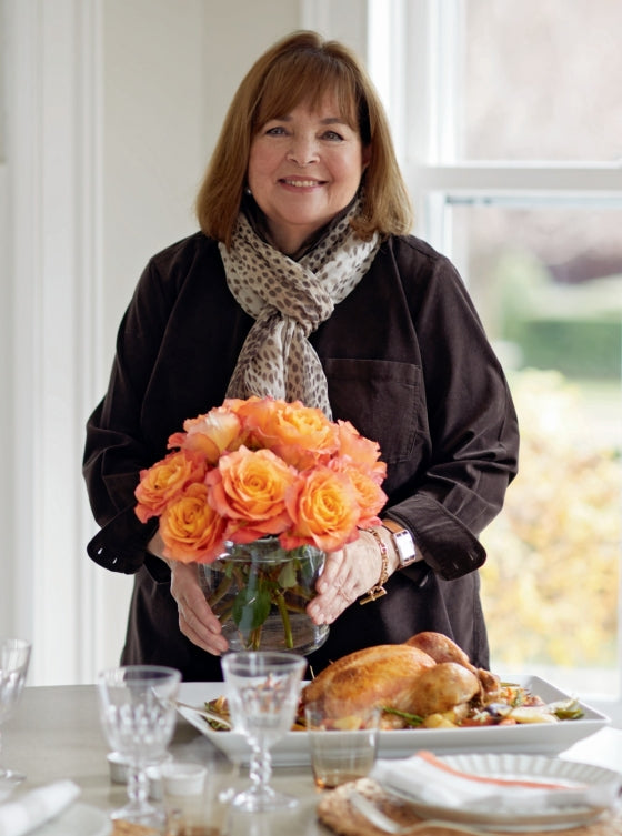 A woman stands indoors holding a vase of orange roses behind a table set with dishes, glassware, and a beautifully roasted chicken—the perfect setting to inspire easy dinner ideas or comforting recipes from Go-to Dinners by Penguin Random House.