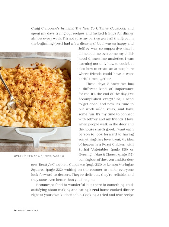 A baked macaroni and cheese dish topped with breadcrumbs sits in a white casserole dish beside a serving spoon on a white background, making it the perfect choice for fans of comforting recipes and easy dinner ideas featured in *Go-to Dinners* from Penguin Random House.