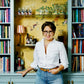 A person in a white shirt and jeans stands in front of built-in bookshelves filled with colorful books, including “Having People Over” by Penguin Random House, beside a small bar area perfect for entertaining or preparing a charcuterie board.