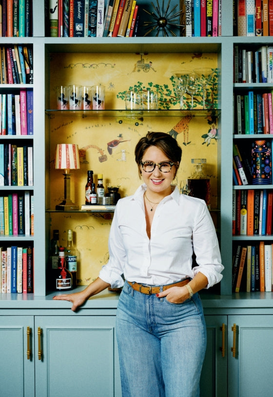 A person in a white shirt and jeans stands in front of built-in bookshelves filled with colorful books, including “Having People Over” by Penguin Random House, beside a small bar area perfect for entertaining or preparing a charcuterie board.