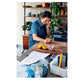 A person stands in their kitchen beside a wooden counter strewn with lemons, papers, and utensils—perhaps paging through Ottolenghi Comfort by Penguin Random House—as they touch their face and smile. In the background, shelves lined with plants and dishes enhance the cozy, comforting cooking atmosphere.