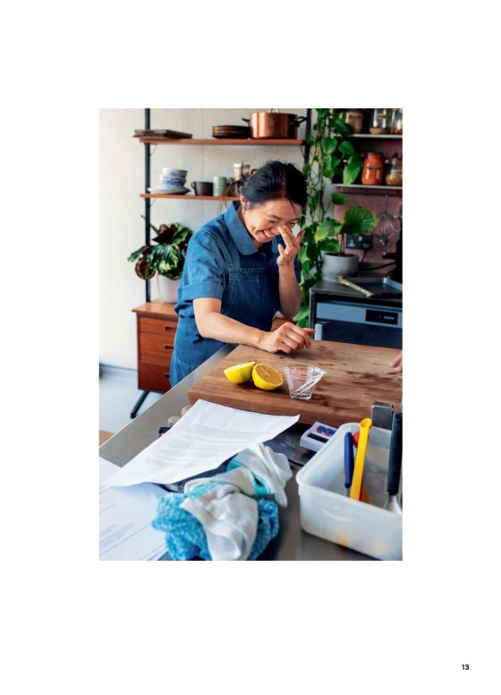 A person stands in their kitchen beside a wooden counter strewn with lemons, papers, and utensils—perhaps paging through Ottolenghi Comfort by Penguin Random House—as they touch their face and smile. In the background, shelves lined with plants and dishes enhance the cozy, comforting cooking atmosphere.