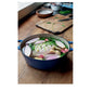 A raw whole chicken rests in a blue pot alongside shallots, garlic, and herbs on a wooden table with a cutting board and bowls nearby, capturing the cozy essence of home cooking inspired by Ottolenghi Comfort from Penguin Random House.