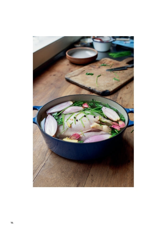A raw whole chicken rests in a blue pot alongside shallots, garlic, and herbs on a wooden table with a cutting board and bowls nearby, capturing the cozy essence of home cooking inspired by Ottolenghi Comfort from Penguin Random House.