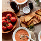 A cozy spread featuring two bowls of tomato soup with herbs, grilled cheese sandwiches, a wooden bowl of tomatoes, a wine bottle, a glass of white wine, salt, and a pepper grinder sits on a rustic wooden table—evoking the delicious inspiration found in Modern Comfort Food by Ina Garten from Penguin Random House. Ideal for anyone who loves comforting home-cooked recipes.