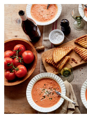 A cozy spread featuring two bowls of tomato soup with herbs, grilled cheese sandwiches, a wooden bowl of tomatoes, a wine bottle, a glass of white wine, salt, and a pepper grinder sits on a rustic wooden table—evoking the delicious inspiration found in Modern Comfort Food by Ina Garten from Penguin Random House. Ideal for anyone who loves comforting home-cooked recipes.