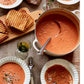 A pot of tomato soup with a ladle, three bowls of served soup, grilled bread slices on a wooden board, and glasses of white wine on a rustic table—just the kind of inviting scene you’ll discover in Modern Comfort Food by Ina Garten, published by Penguin Random House, where classic comfort food recipes truly come to life.