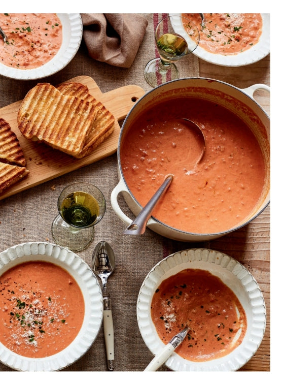 A pot of tomato soup with a ladle, three bowls of served soup, grilled bread slices on a wooden board, and glasses of white wine on a rustic table—just the kind of inviting scene you’ll discover in Modern Comfort Food by Ina Garten, published by Penguin Random House, where classic comfort food recipes truly come to life.