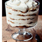 A glass trifle dish filled with layered cookies, cream, and banana slices sits on a wooden surface next to a knife and some broken cookies—a picture-perfect treat inspired by recipes from Modern Comfort Food by Ina Garten, published by Penguin Random House.