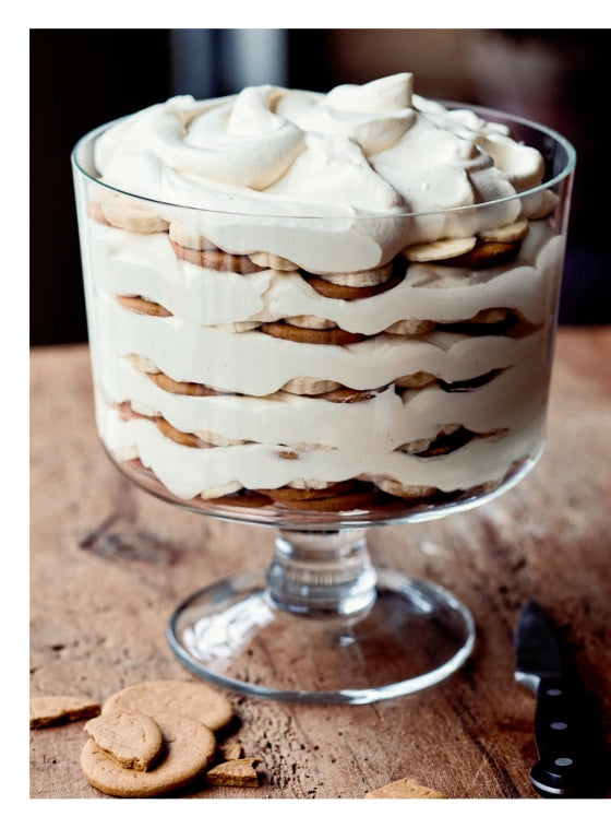 A glass trifle dish filled with layered cookies, cream, and banana slices sits on a wooden surface next to a knife and some broken cookies—a picture-perfect treat inspired by recipes from Modern Comfort Food by Ina Garten, published by Penguin Random House.