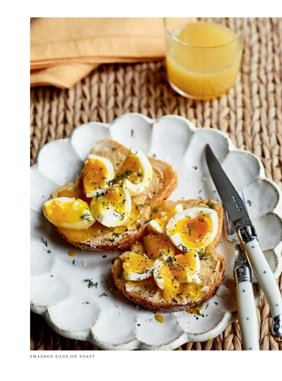 Two slices of toast topped with sliced soft-boiled eggs and herbs grace a white plate, accompanied by a knife, fork, and a glass of orange juice—an inviting scene inspired by recipes from Modern Comfort Food by Ina Garten, published by Penguin Random House, bringing warmth and simplicity to your breakfast table.