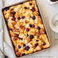 A rectangular baking dish filled with bread pudding topped with mixed berries sits on a light surface next to a spoon, a napkin, a bowl of sugar, and a small bowl of sauce—an inviting scene inspired by recipes from The Couple’s Cookbook by Penguin Random House, perfect for couples who love cooking together.