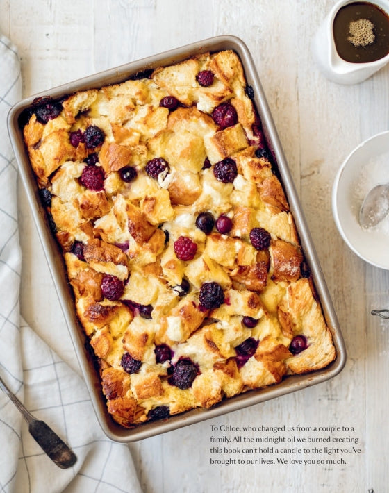 A rectangular baking dish filled with bread pudding topped with mixed berries sits on a light surface next to a spoon, a napkin, a bowl of sugar, and a small bowl of sauce—an inviting scene inspired by recipes from The Couple’s Cookbook by Penguin Random House, perfect for couples who love cooking together.