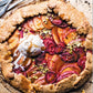 A rustic fruit galette topped with vanilla ice cream, caramel drizzle, and chopped pistachios rests on parchment paper, served comfort-food style with spoons close at hand—a scene inspired by the cozy signature touch of Tieghan Gerard as featured in the Half Baked Harvest Cookbook by Penguin Random House.
